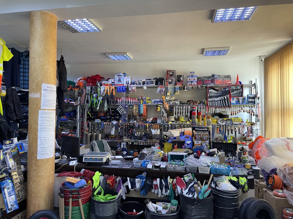 Na zdjęciu widać obszerną ladę sklepowa wypełnioną różnorodnymi produktami, w tym koszykami z drobnymi akcesoriami, taśmami, rękawicami, narzędziami oraz elektroniką, za którą stoją wagi, terminal i stanowisko kasowe. W tle znajduje się ściana z metalowymi regałami, na których zawieszone są klucze, wkrętaki, piły, tarcze szlifierskie, smary, spraye i liczne akcesoria techniczne oraz ogrodnicze. Przed ladą, na dole, widoczne są kolorowe wiadra, węże ogrodowe, rękawice robocze i inne wyposażenie, a po bokach fragmenty odzieży roboczej i materiałów opakowaniowych, co podkreśla szeroki asortyment sklepu.