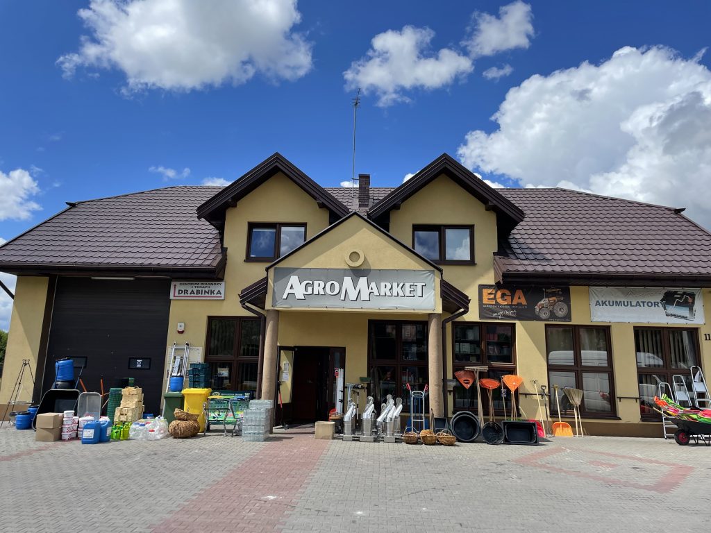 Na zdjęciu widać żółty, dwukondygnacyjny budynek sklepu AgroMarket z brązowym, spadzistym dachem i dużym szyldem nad wejściem głównym. Przed sklepem ustawiono różne narzędzia i akcesoria rolniczo‑ogrodnicze, takie jak taczki, łopaty, grabie, miotły, kosze wiklinowe, plastikowe pojemniki i kanistry, a także metalowe urządzenia stojące w rzędzie. Po prawej stronie widoczne są banery reklamowe, a brukowany podjazd prowadzi bezpośrednio do otwartych drzwi sklepu, wszystko w jasnym, słonecznym świetle z kilkoma białymi chmurami na niebie.