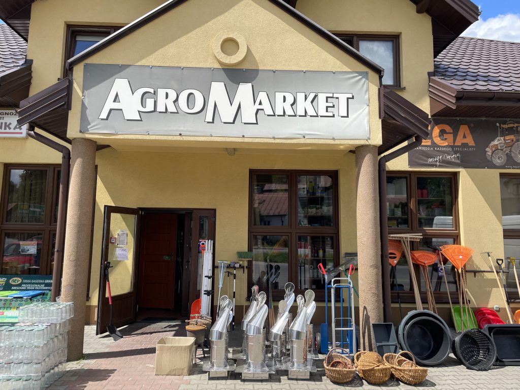 Wejście do sklepu AgroMarket z narzędziami ogrodniczymi i sprzętem rolniczym przed budynkiem.